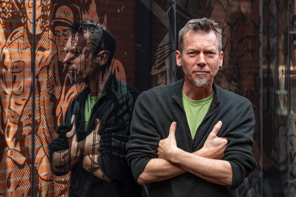 ein Mann lächelt in die Kamera, hinter ihm ist die Spiegelung des Kubus zu erkennen