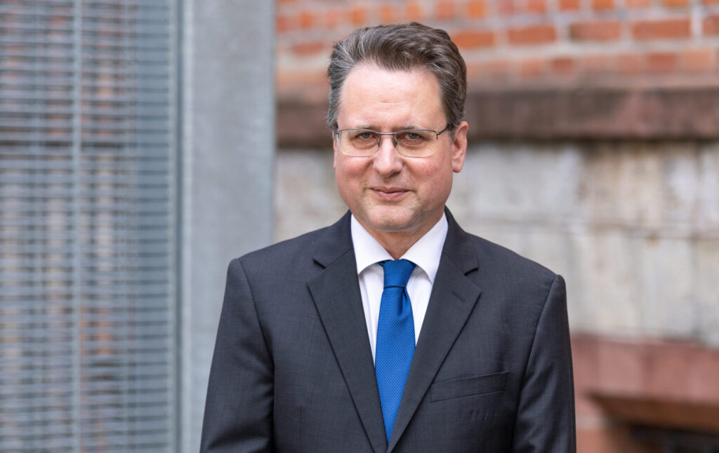 ein Mann lächelt in die Kamera, hinter ihm sind eine rote Backsteinwand und eingraues Gitter zu sehen