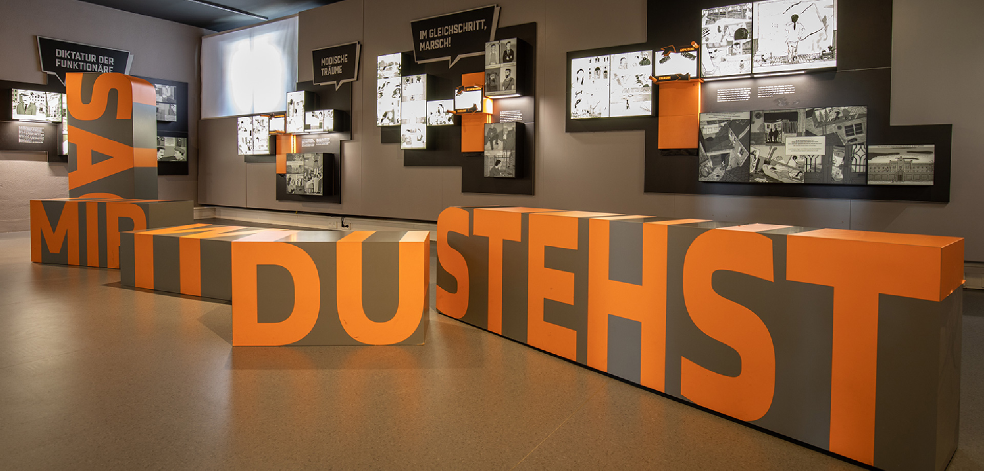 ein Ausstellungsraum: An der Wand sind beleuchtete Kästen angebracht, im Raum stehen große Kästen, auf diesen ist zu lesen: "Sag mir wo du stehst"