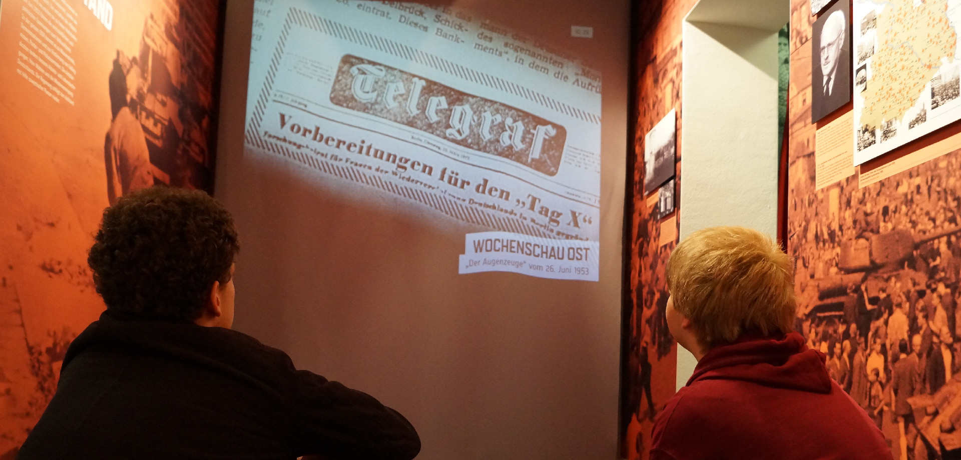 die Hinterköpfe von zwei jungen Mneschen, sie schauen auf eine Leinwand mit historischen Aufnahmen. Zu sehen ist der Ausschnitt einer Zeitung, darauf steht "Telegraf. Vorbereitungen für den Tag X", darunter der Hinweis auf "Wochenschau Ost"