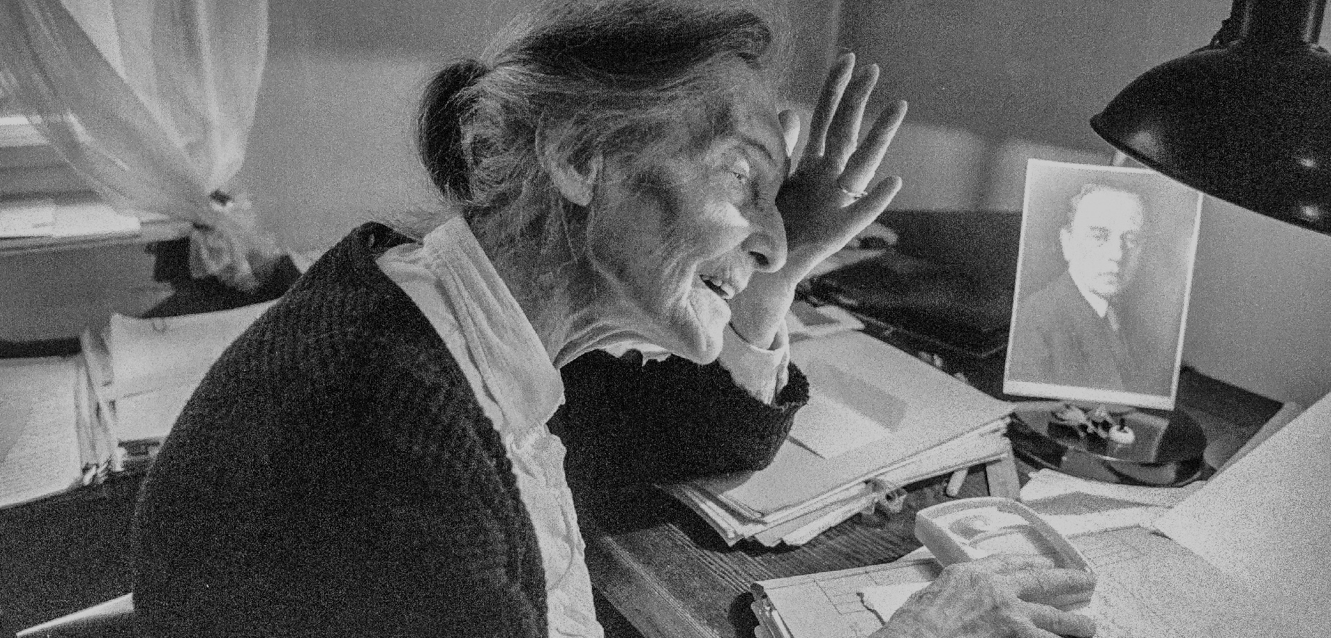 eine alte Frau sitzt an einem Schreibtisch, auf dem Tisch steht ein Foto von einem jungen Mann, sie hält ihre Hand an ihren Kopf, in der anderen Hand hat sie eine Lupe, mit der liest sie in einem Dokument