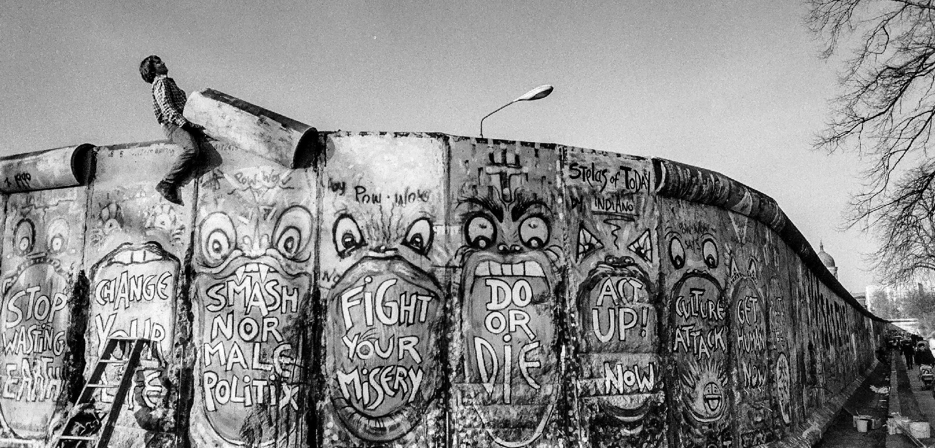 eine Mauer aus mehreren Platten, die Mauer ist mit Gesichtern und Sprüchen vollgemalt. Ein junger MAnn sitzt auf der MAuer und hebt einen schweren Verbidungsstein runter.
