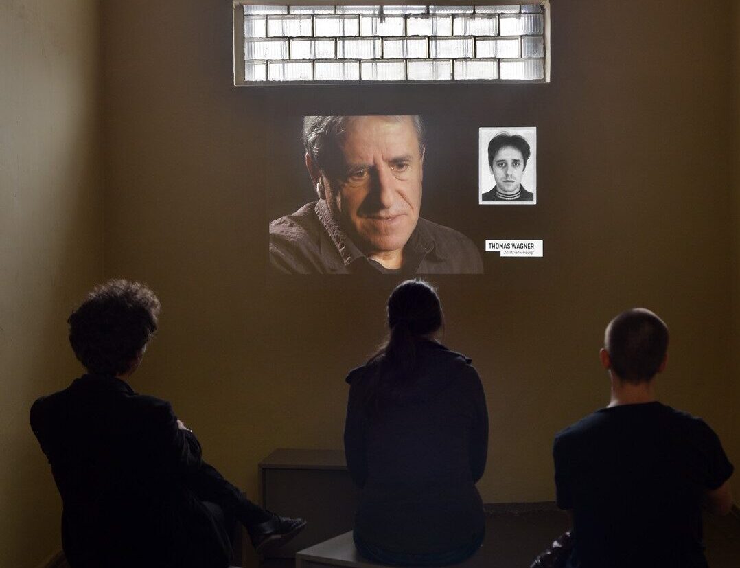 drei Menschen sitzen vor einer Wand, auf der ein Mann zusehen ist, darüber ist ein Fenster aus Glasbausteinen