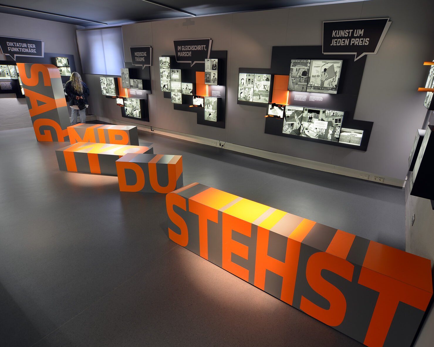 Blick in einen Raum: An der Wand sind leuchtende Kästen angebracht, in der Mitte sind Aufsteller, auf denen ist zu lesen: 