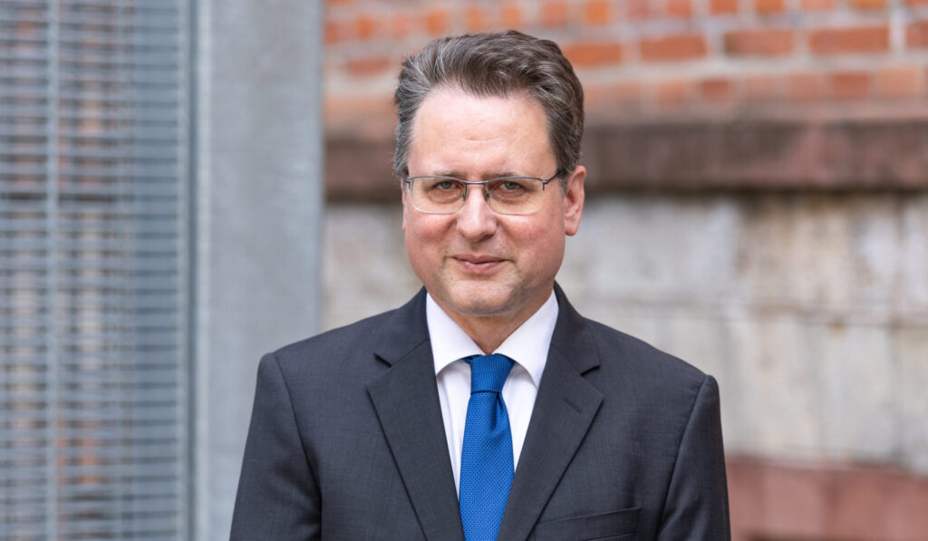 ein Mann lächelt in die Kamera, hinter ihm sind eine rote Backsteinwand und eingraues Gitter zu sehen