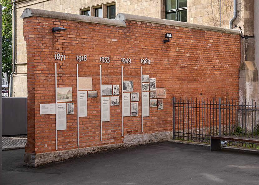 Eine Backsteinmauer mit Tafeln und verschiedenen Jahreszahlen zwischen 1871 und 1989 darauf