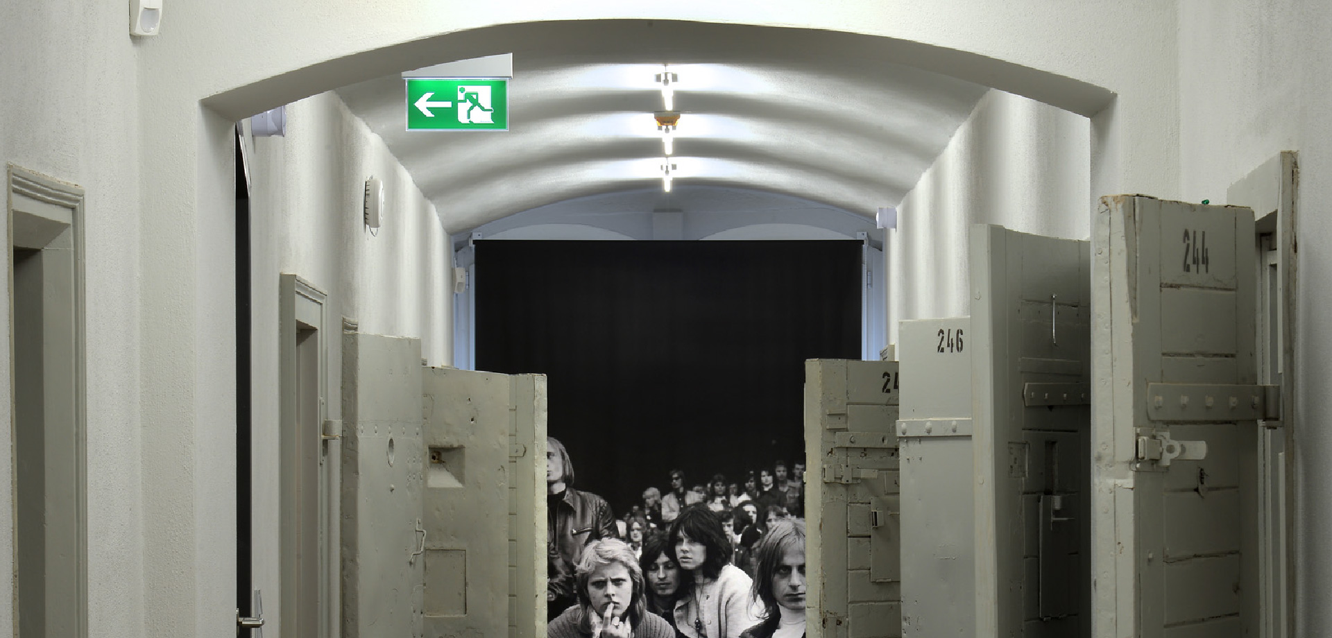 Ein Gang mit offenen Hafttüren, im Hintergrund ist ein Foto in schwarz/weiß mit jungen Menschen zu sehen