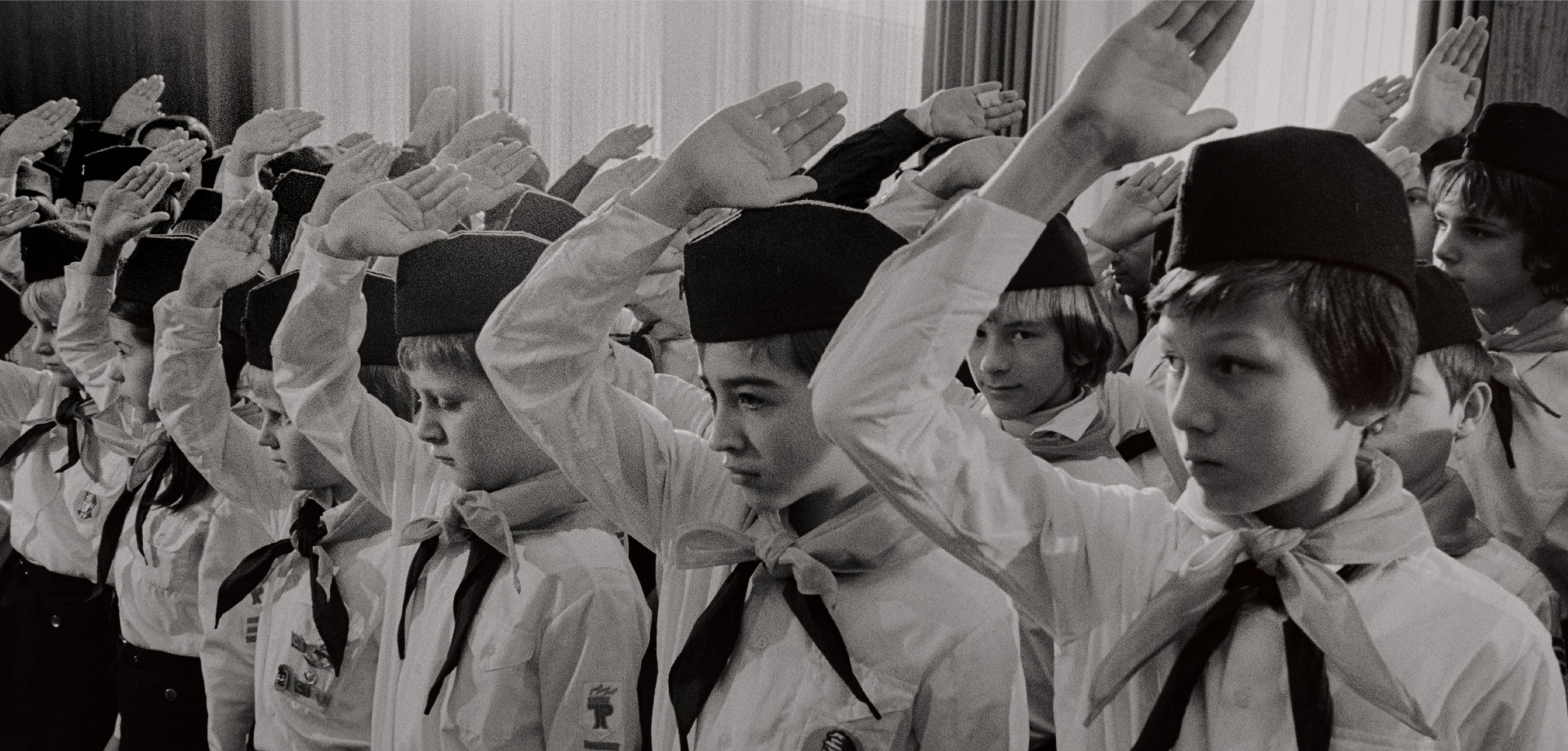 junge Mneschen stehen in mehreren Reihen. Sie tragen weiße Hemden, ein Halstuch und Hüte. Sie haben alle die rechte Hand parallel zum Kopf gehoben