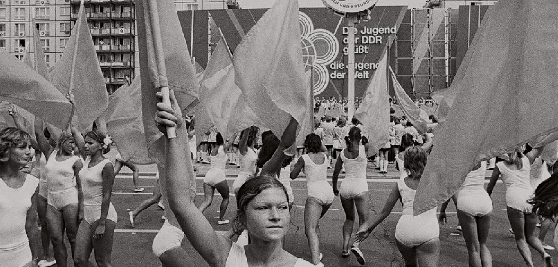 viele Frauen in weißen Turnanzügen und Fahnen, die auf einem grßen Platz tanzen. Im Hintergrund steht "Die JUgend der DDR grüßt die Jugen der Welt"