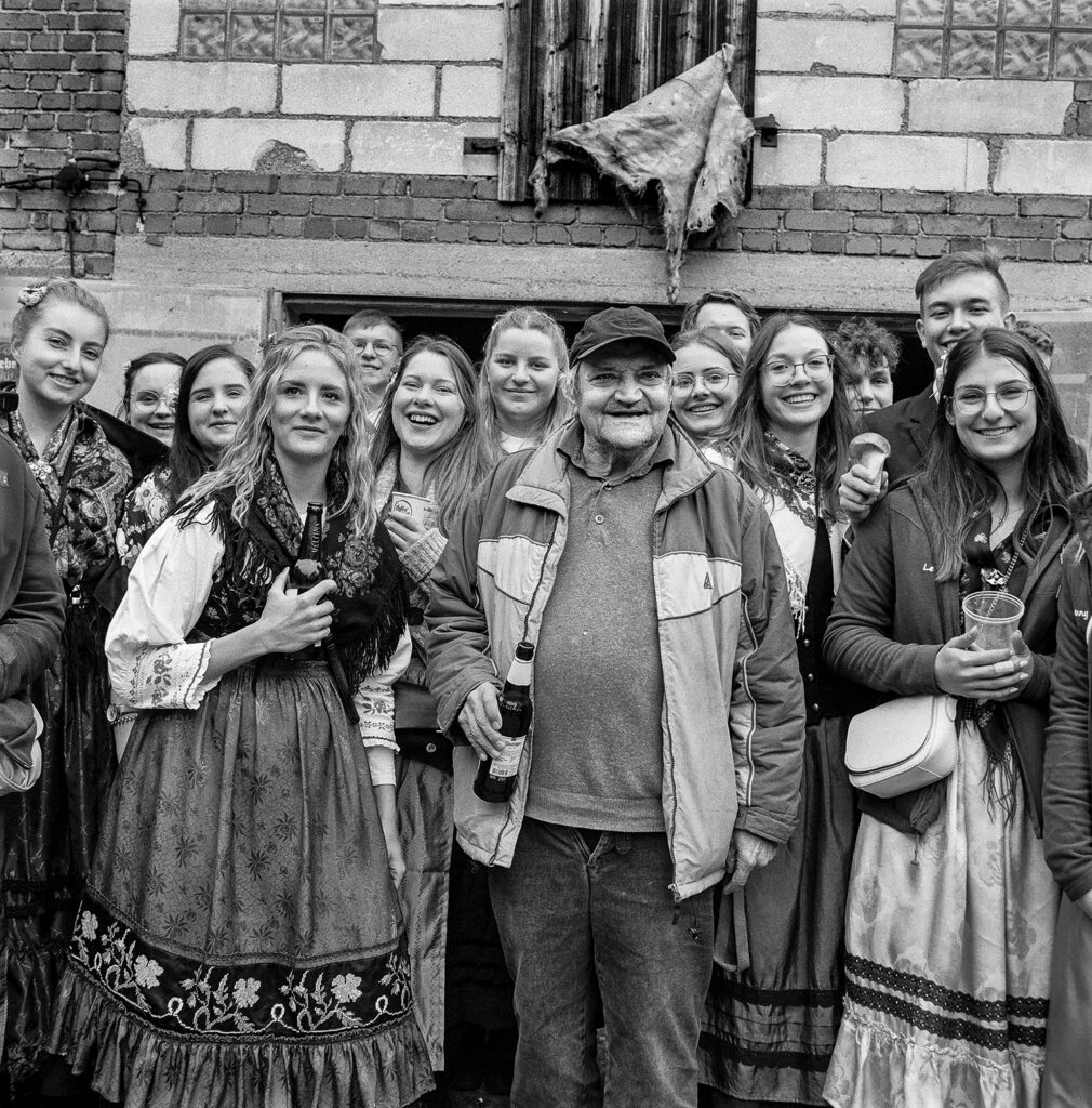 Ein älterer Mann steht in einer Gruppe junger Menschen, hauptsächlich Frauen. Der Mann hat eine Bierflschae in der Hand und trägt eine Sportjacke. Er lächelt in die Kamera. Die jugen Frauen lächeln ebenfalls in die KAmera. Sie tragen Trachten und auch sie haben Flaschen oder Becher in den Händen.