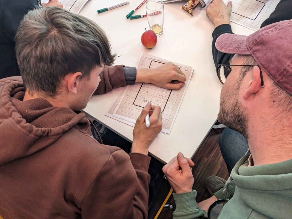 Blick von oben auf zwei junge Männer, man sieht ihre Hinterkoöpfe; Sie beugen sich über ein Blatt, einer der beiden zeichnet etwas in eine vorgegebene Form, der andere schaut ihm dabei zu.